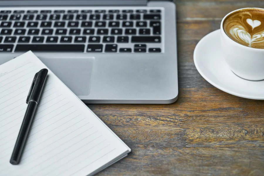 A laptop, an open notebook with a pen, and a coffee on a desk