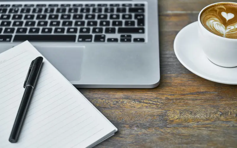 A laptop, an open notebook with a pen, and a coffee on a desk